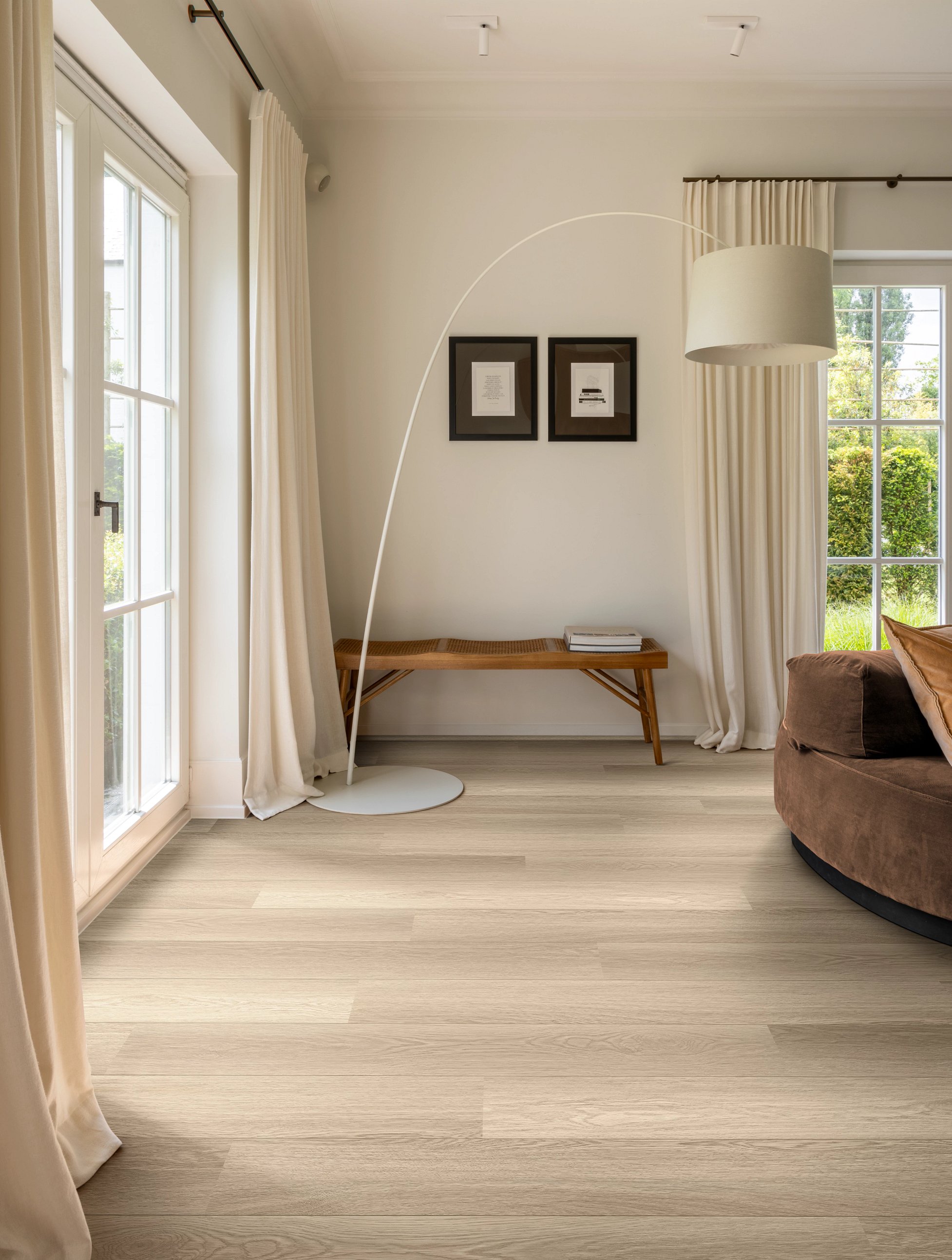 Ein gemütlich eingerichtetes Wohnzimmer mit einem beigen Sofa, Holzboden, einer modernen Stehlampe und großen Fenstern mit Vorhängen.