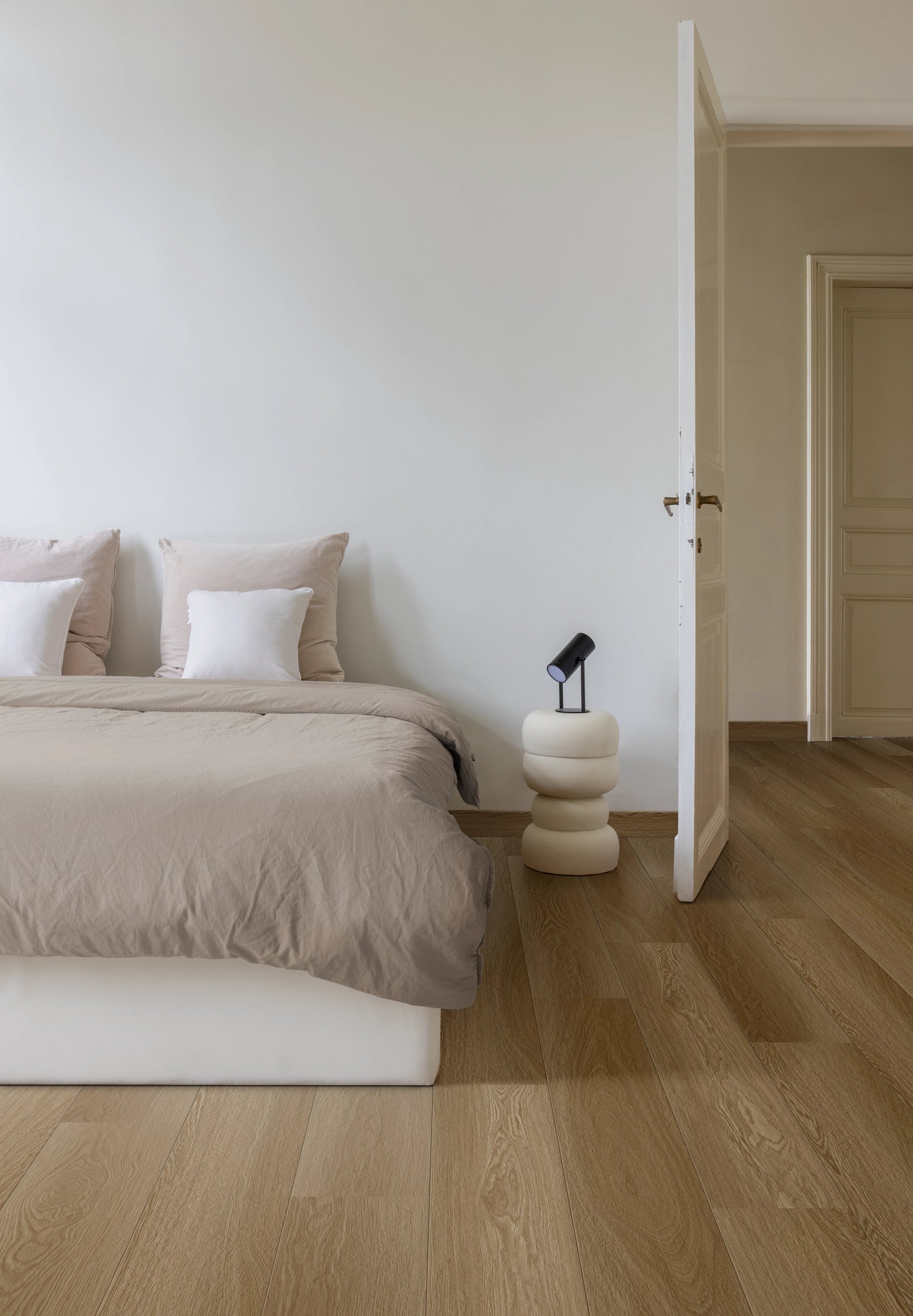 Ein modernes Schlafzimmer mit einem Bett in beige Bettwäsche neben einem weißen, gestapelten Nachttisch mit schwarzer Lampe, Holzboden und offener Tür.