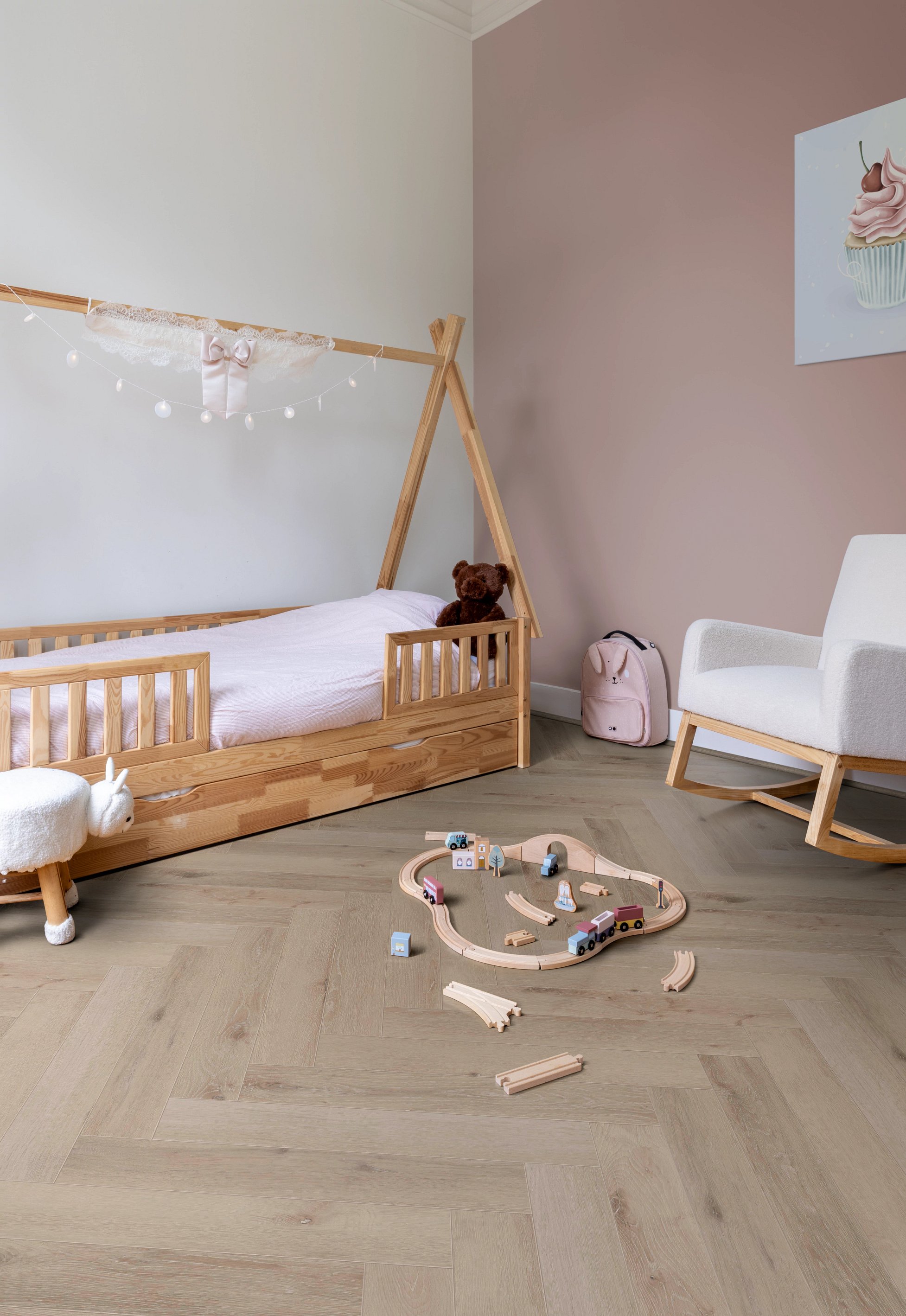 Ein Kinderzimmer mit einem Holzhausbett, Kuscheltier, Schaukelstuhl und einer auf dem Boden aufgebauten Spielzeugeisenbahn.